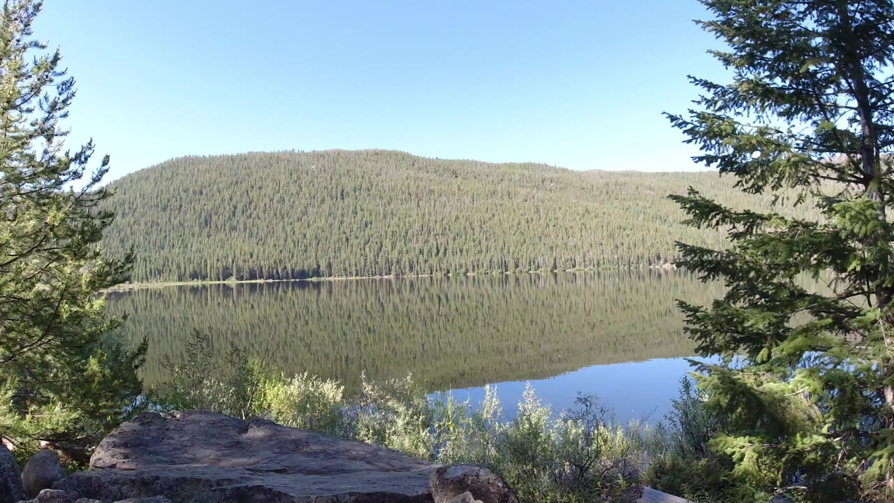 Winter Park, Colorado lake view from a rocky overlook, with tall pines at the edges, forested hills across the water, bright blue sky, and no visible people.