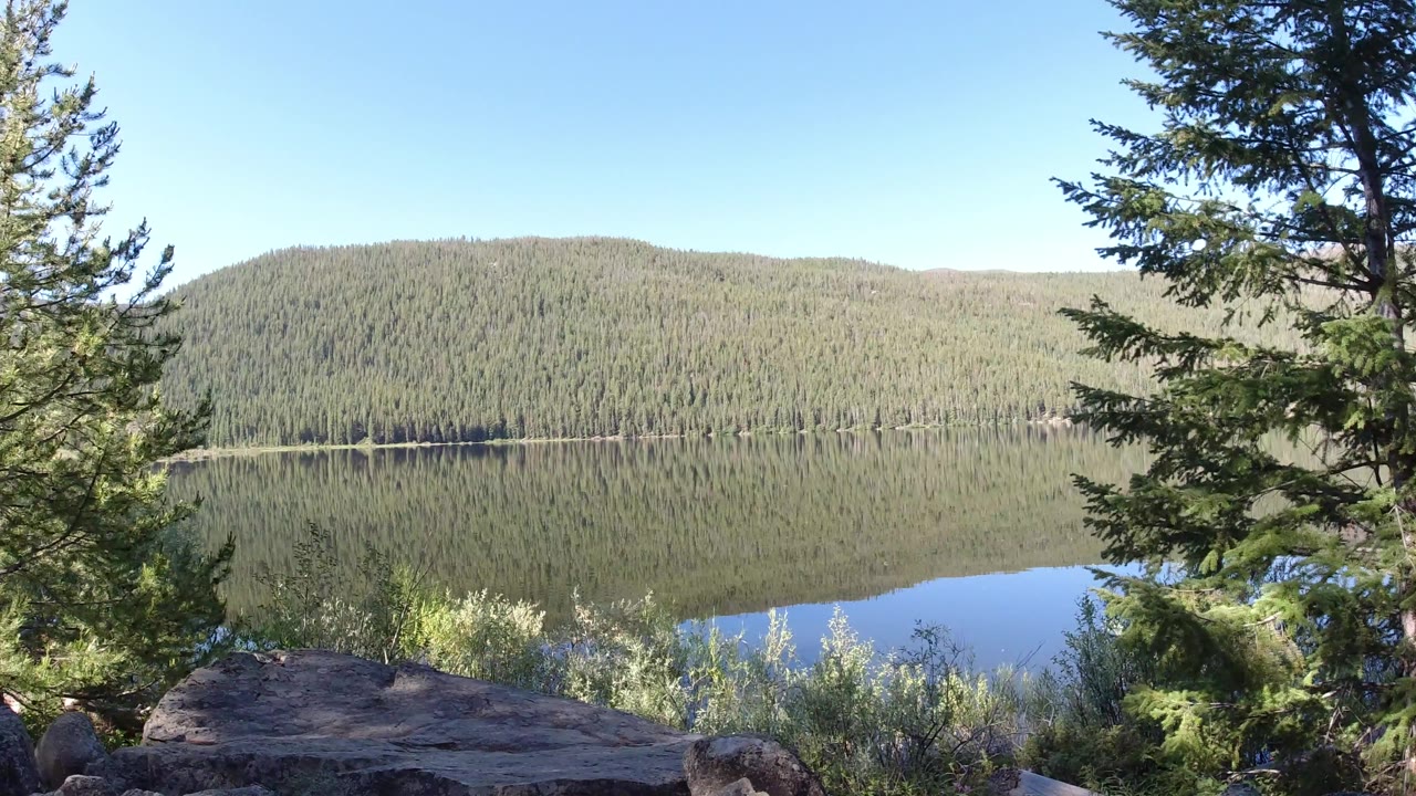 Winter Park, Colorado overlook with a broad rock ledge in front, evergreen branches crossing the center, a still lake below, wooded hills beyond, and no visible people.