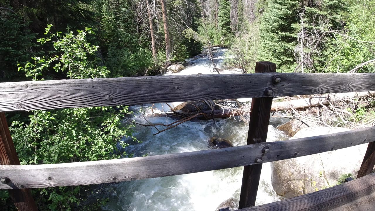 At 0:00, weathered bridge rails cross the foreground above white river rapids and green forest in Winter Park, Colorado, with no visible people.