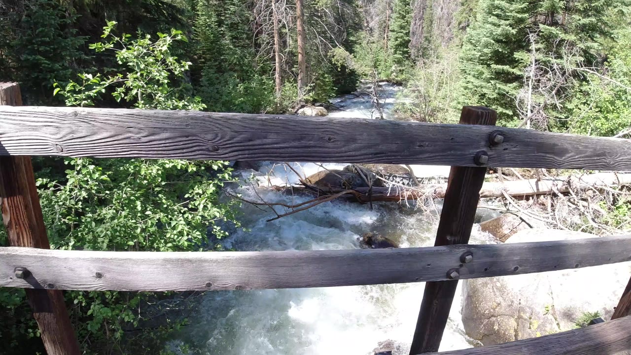 Weathered wooden bridge railing crosses a view of a rushing stream and green trees in Winter Park, Colorado, with no visible people.