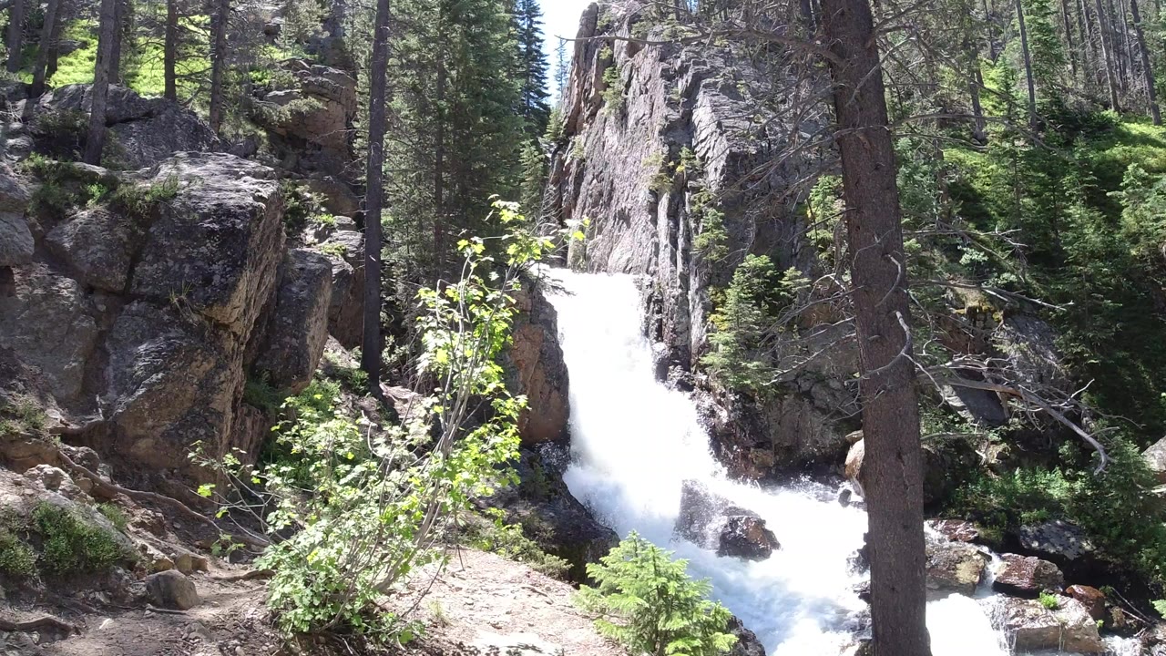 A bright waterfall drops between gray rocks and conifer trees in Winter Park, Colorado, with green shrubs in the foreground and no visible people.