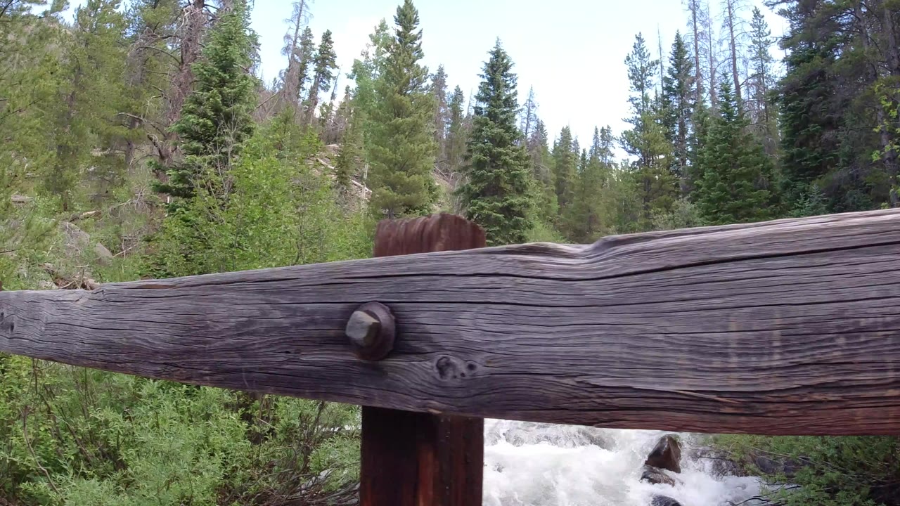 Weathered wooden bridge rail crosses the foreground in Winter Park, Colorado, with white creek water and tall green forest behind it, no visible people.