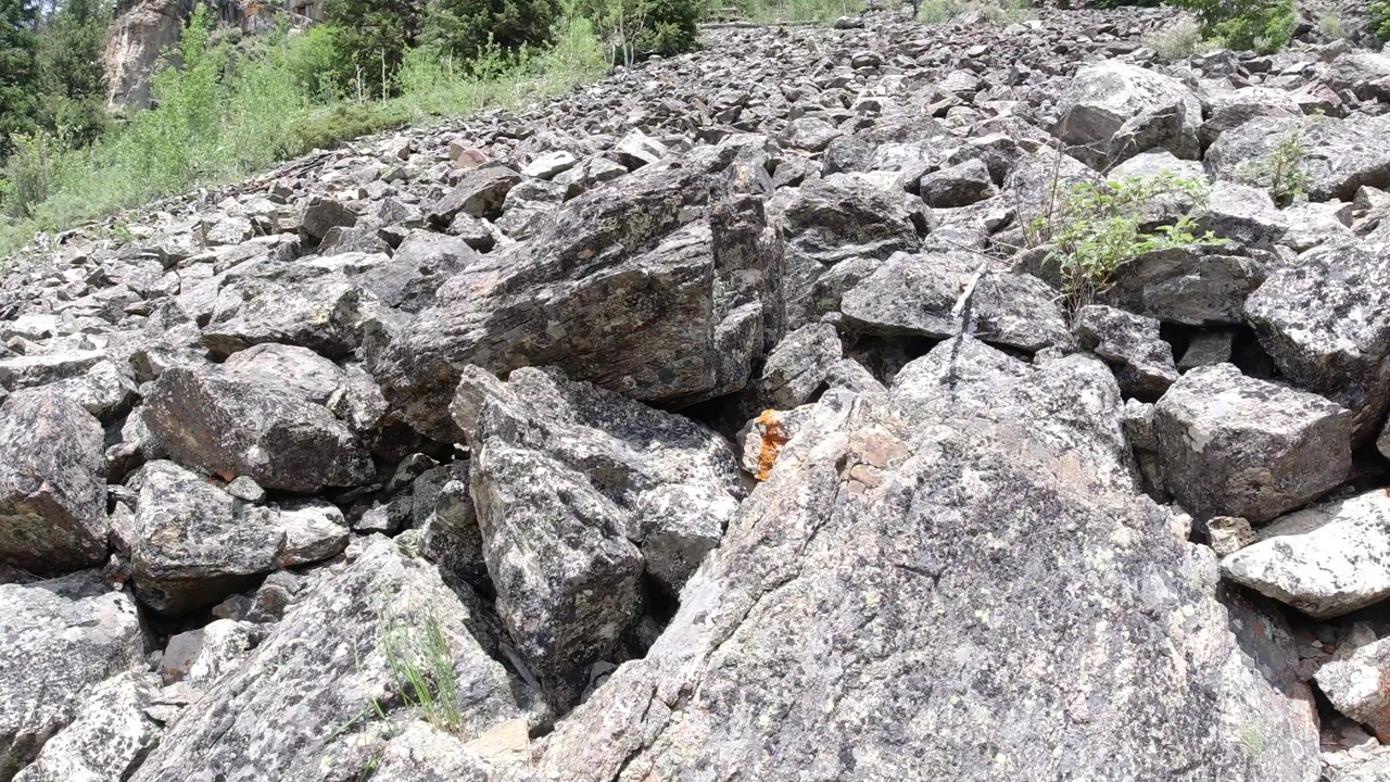 Start frame of a rocky slope in Winter Park, Colorado, with large gray boulders in front, smaller stones uphill, green trees at the top edge, and no visible people.