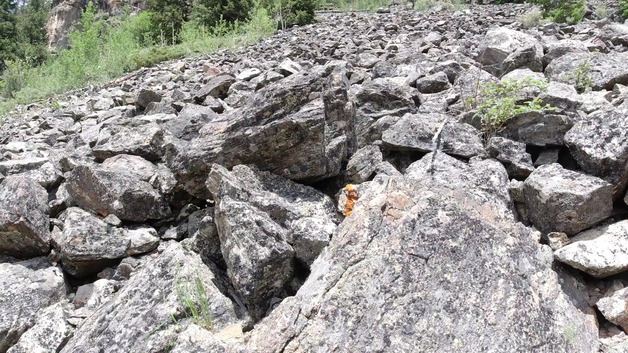 Rock-lined terrain in Winter Park, Colorado, with a broad foreground boulder, broken stones across the center, shrubs near the upper edge, and no visible people.