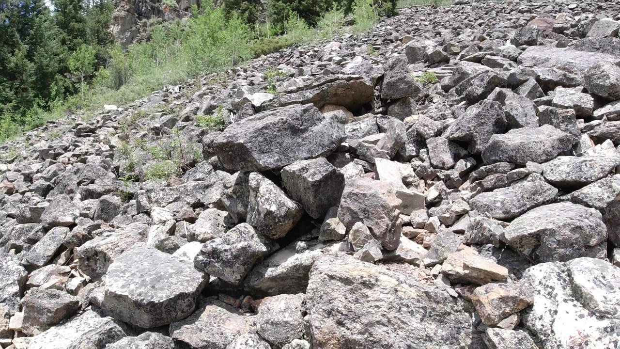 A dense field of gray and tan rocks in Winter Park, Colorado, fills the frame, with a larger flat boulder near the center, scrubby vegetation along the upper slope, and no visible people.