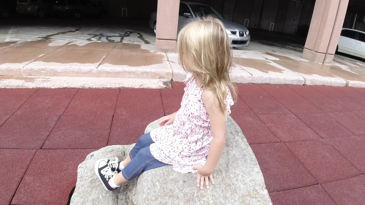 Young blonde girl sitting on a rock beside red sidewalk pavers.