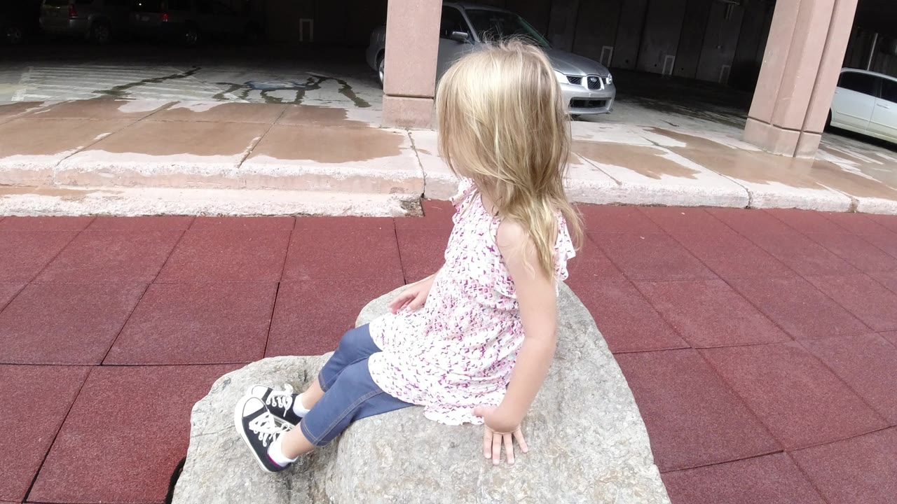 The girl remains seated on the rock in Winter Park, Colorado, with her back and side to camera and a white car visible under the building overhang.