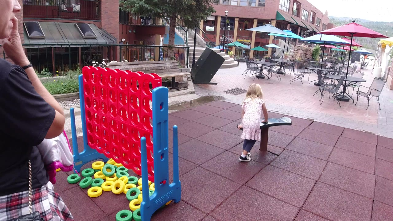 The child walks across the rubber playground surface toward a small green platform, with the red play structure in the foreground and outdoor cafe seating behind her in Winter Park, Colorado.