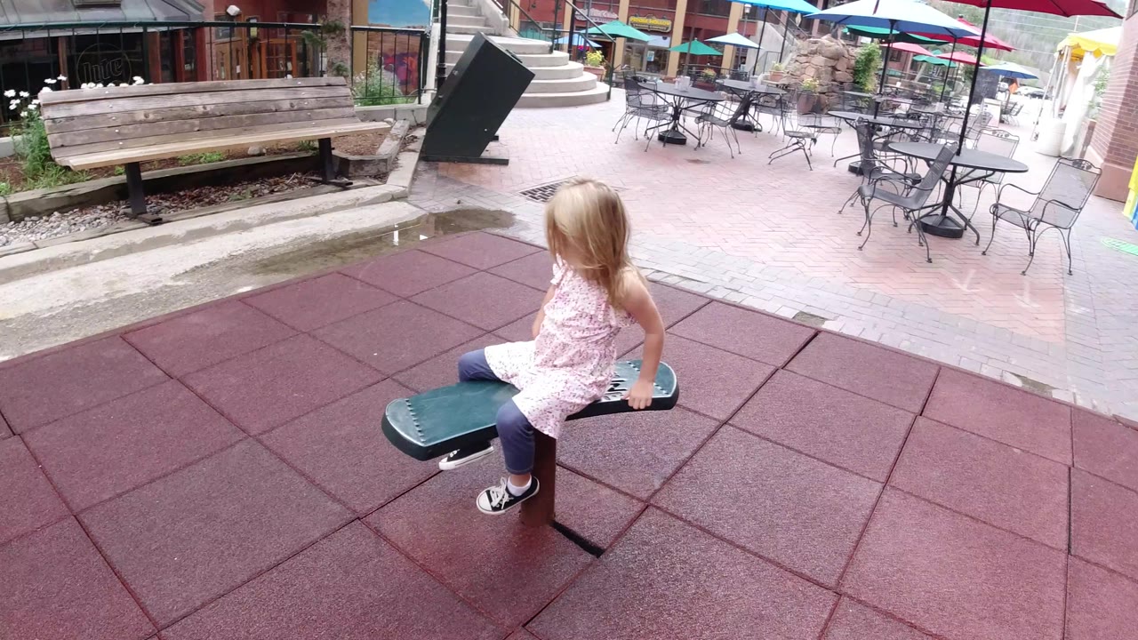 The girl remains seated on the green bouncy seat in the Winter Park outdoor play area, facing toward the patio tables and umbrellas beyond the red tile surface.