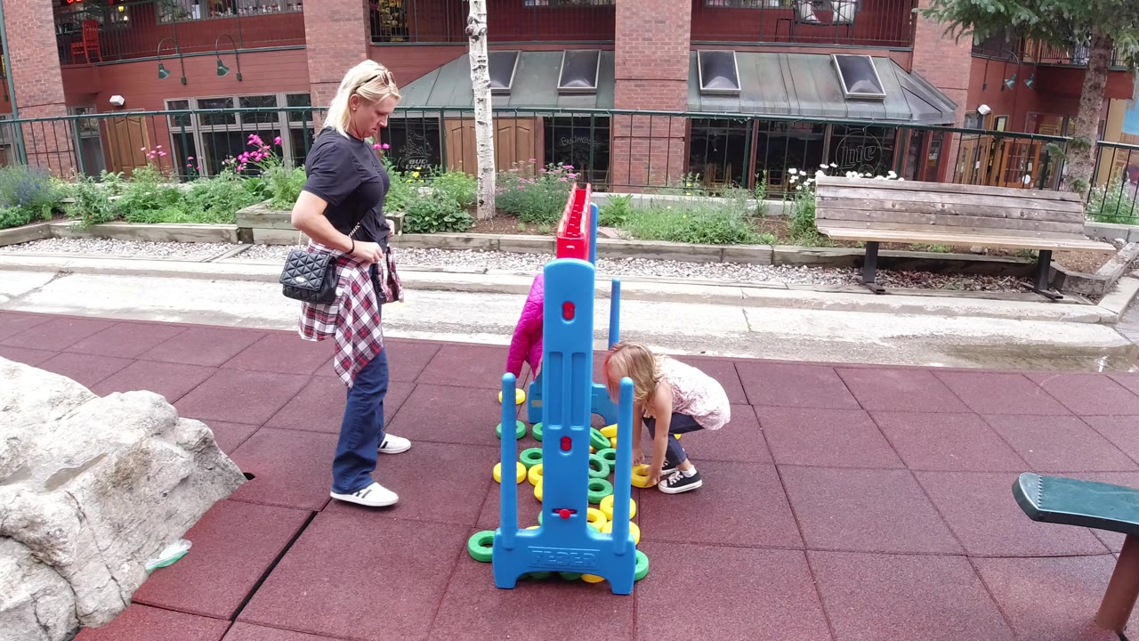Blonde woman watches a young girl play beside a blue playground game.