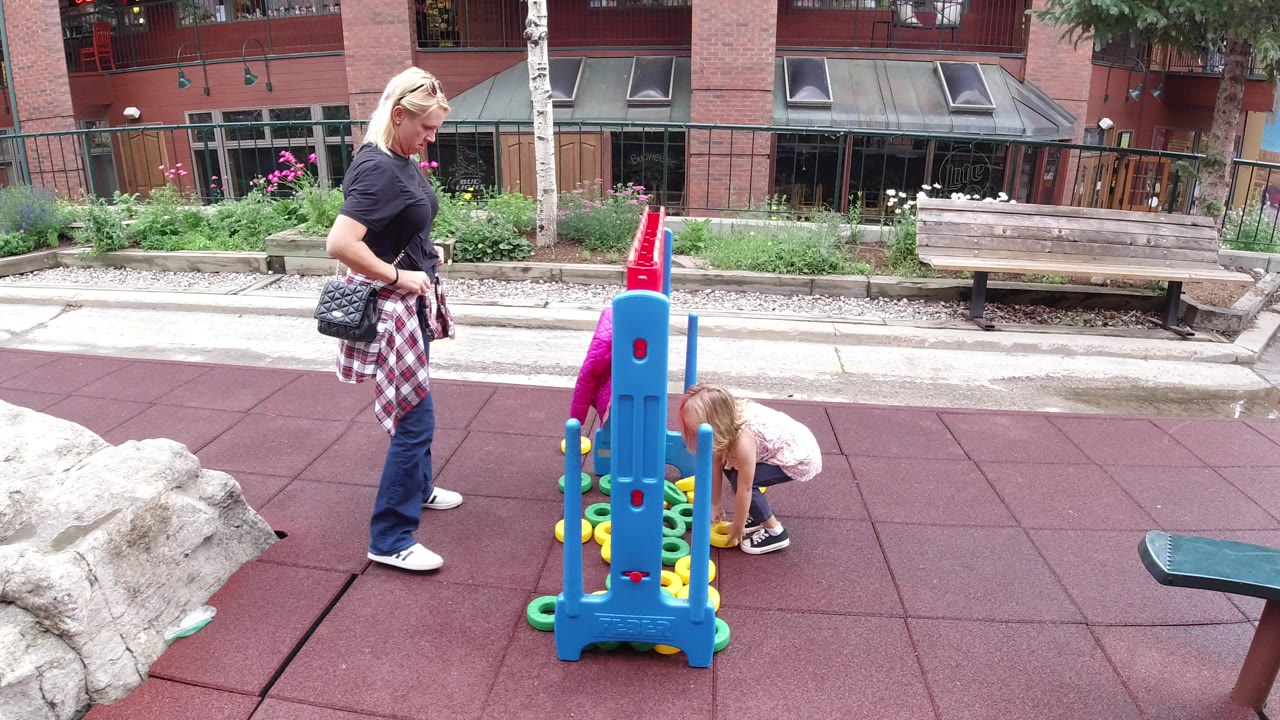 The woman remains beside the blue and red playground structure as the girl reaches down near the scattered play rings on the rubber surface in Winter Park, Colorado.