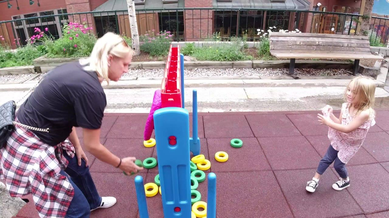 At 0:00 in Winter Park, Colorado, an adult woman bends toward green plastic game pieces while a young blonde girl stands beside a large red-and-blue outdoor game on red rubber tiles.