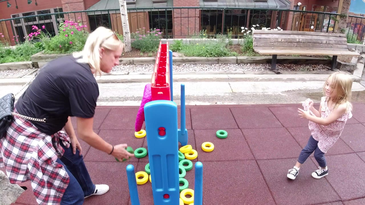 At 0:01 in Winter Park, Colorado, the adult woman remains bent beside the game frame as the young girl faces her, with yellow and green rings scattered around the base.