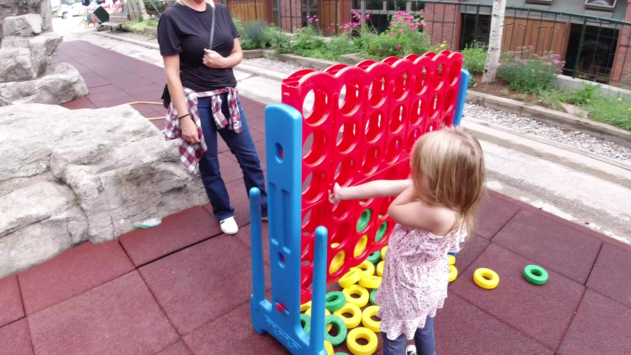 Small girl reaches toward a large red outdoor game while an adult woman stands nearby.
