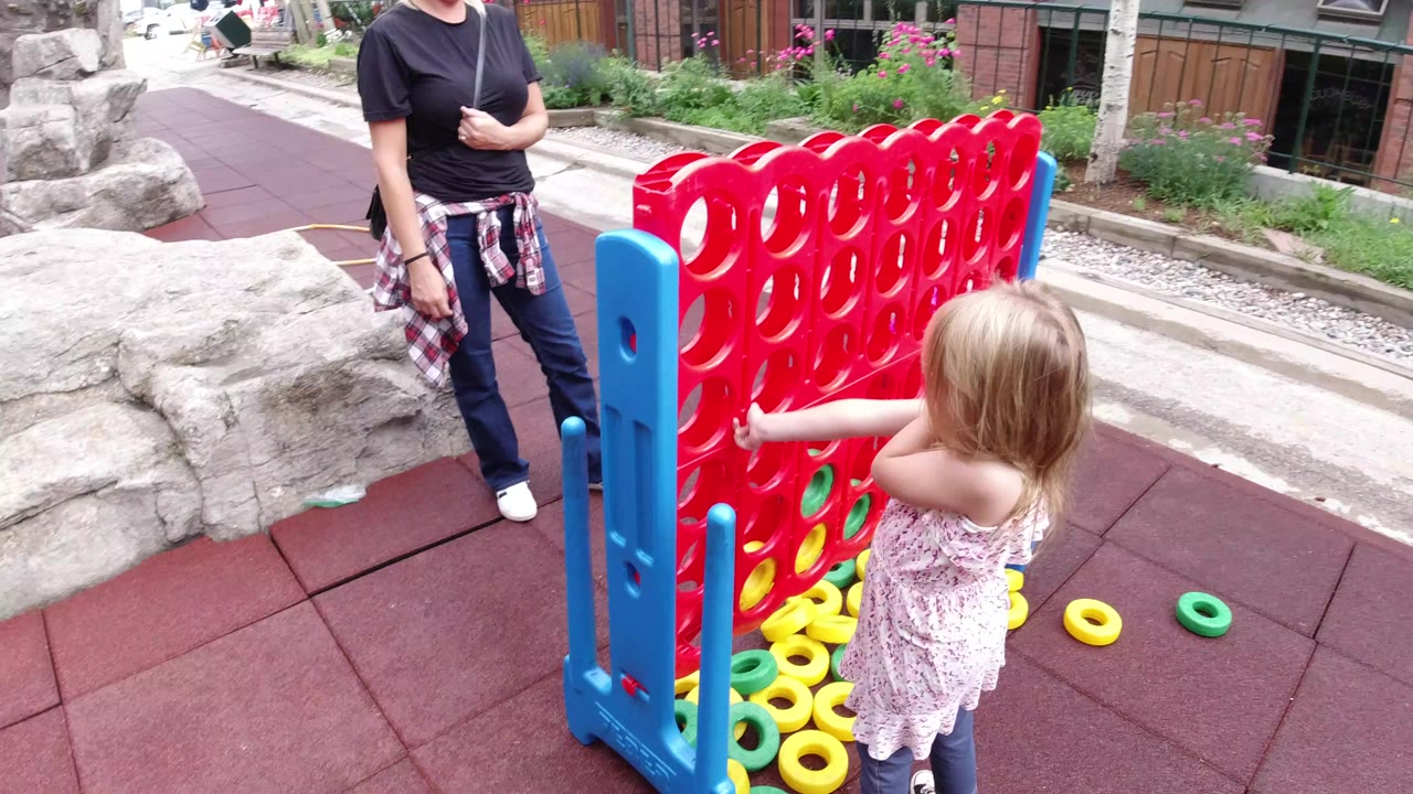 The girl points at the red game board from the right side of frame, with loose yellow and green rings scattered on the red paving and the adult partly visible beside the game.