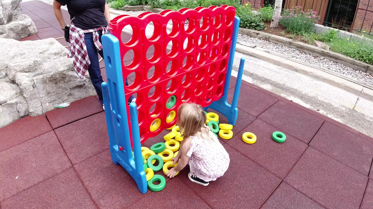 In Winter Park, Colorado, a young girl crouches at the base of a red-and-blue outdoor connect-four style game while an adult woman stands partly in frame nearby.