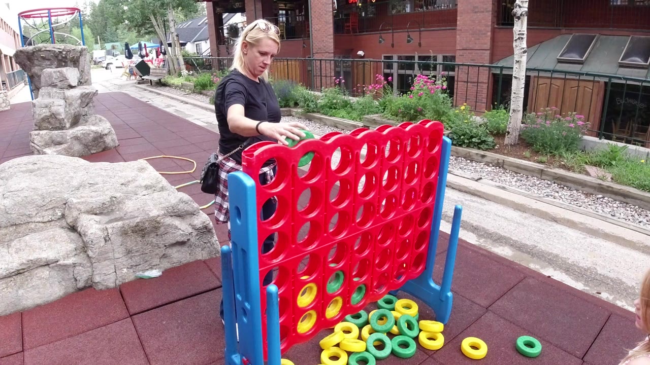 The adult reaches over the red game grid while yellow and green rings remain piled at the base on the outdoor play surface in Winter Park, Colorado.