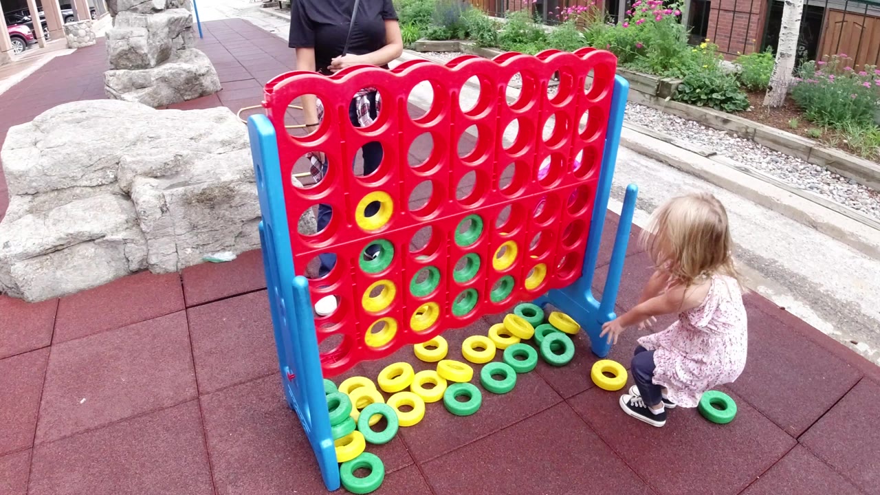 At 0:01 in Winter Park, Colorado, the child remains crouched beside the red game board with loose yellow and green rings lined along the base and one ring near her feet.