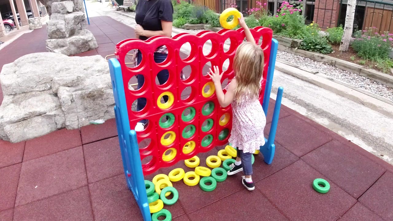 In Winter Park, Colorado, a young girl stands beside a giant red connect-four-style game board while an adult is partly visible behind it.