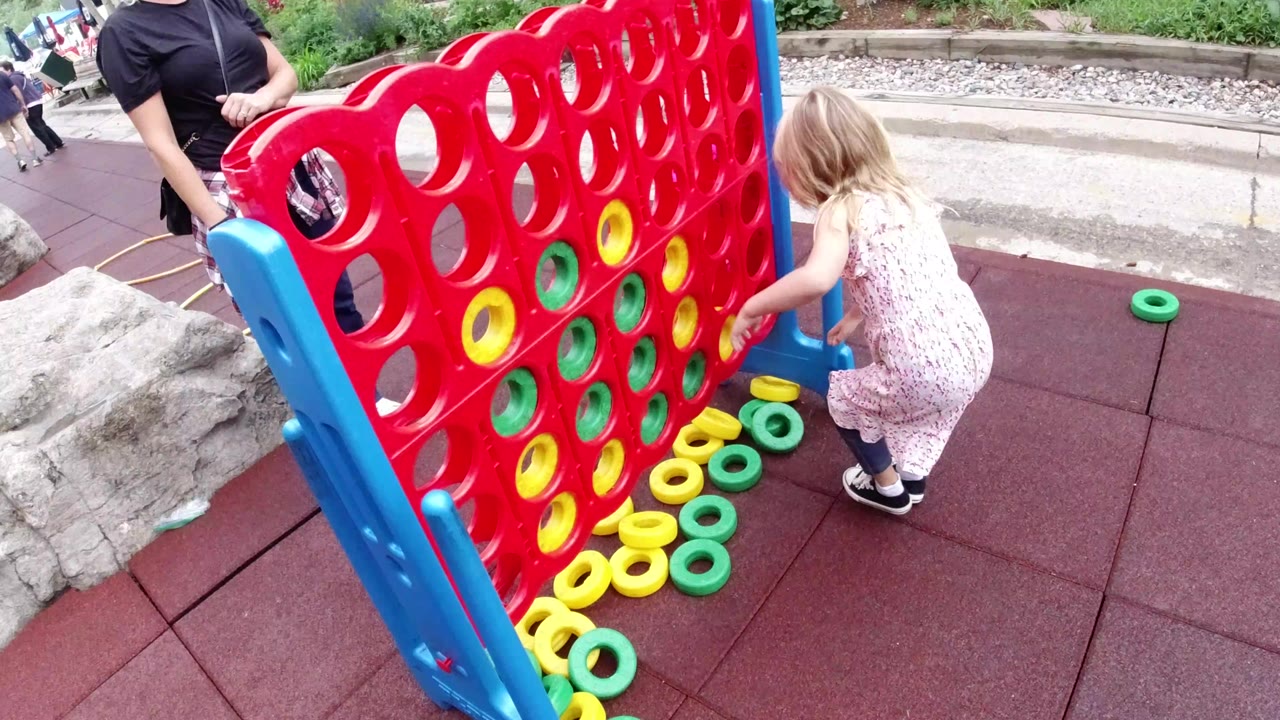 In Winter Park, Colorado, a child crouches at the side of a large red Connect Four style game while yellow and green rings sit on red playground tiles and an adult stands nearby.