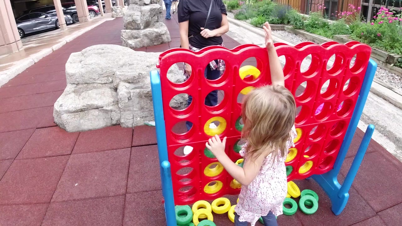 In Winter Park, Colorado, a little blond girl reaches up toward a red outdoor connect-four style game while an adult woman stands partly visible behind it.