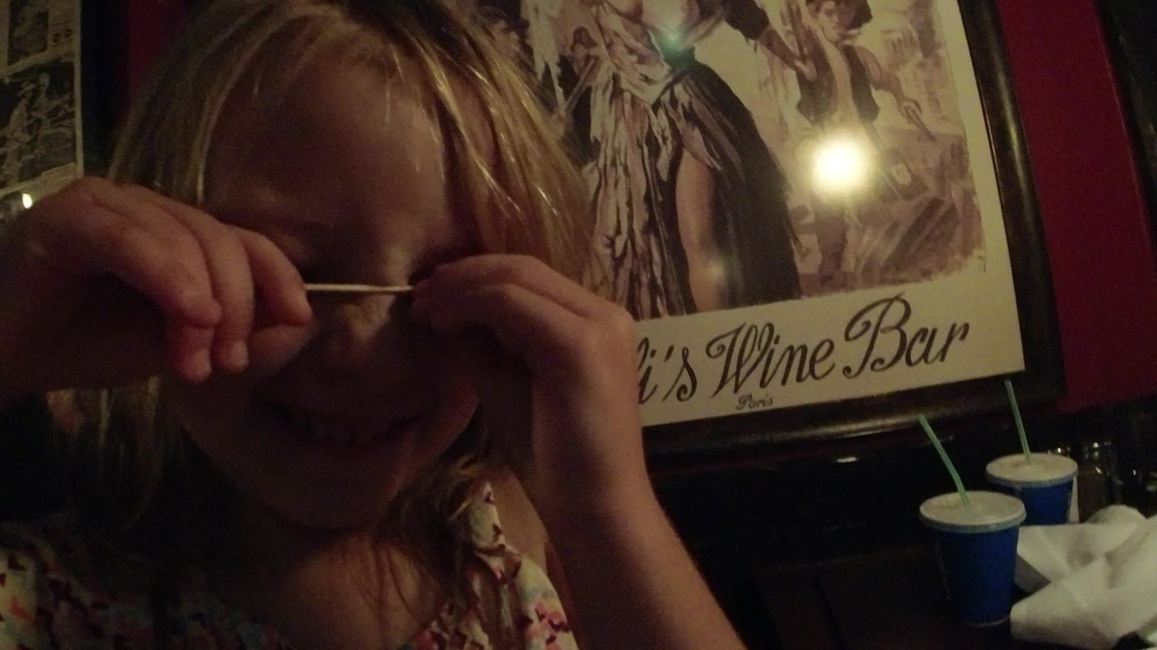 The child smiles in close-up while holding the stick horizontally across her face, with blue cups and framed wall art visible in the background.