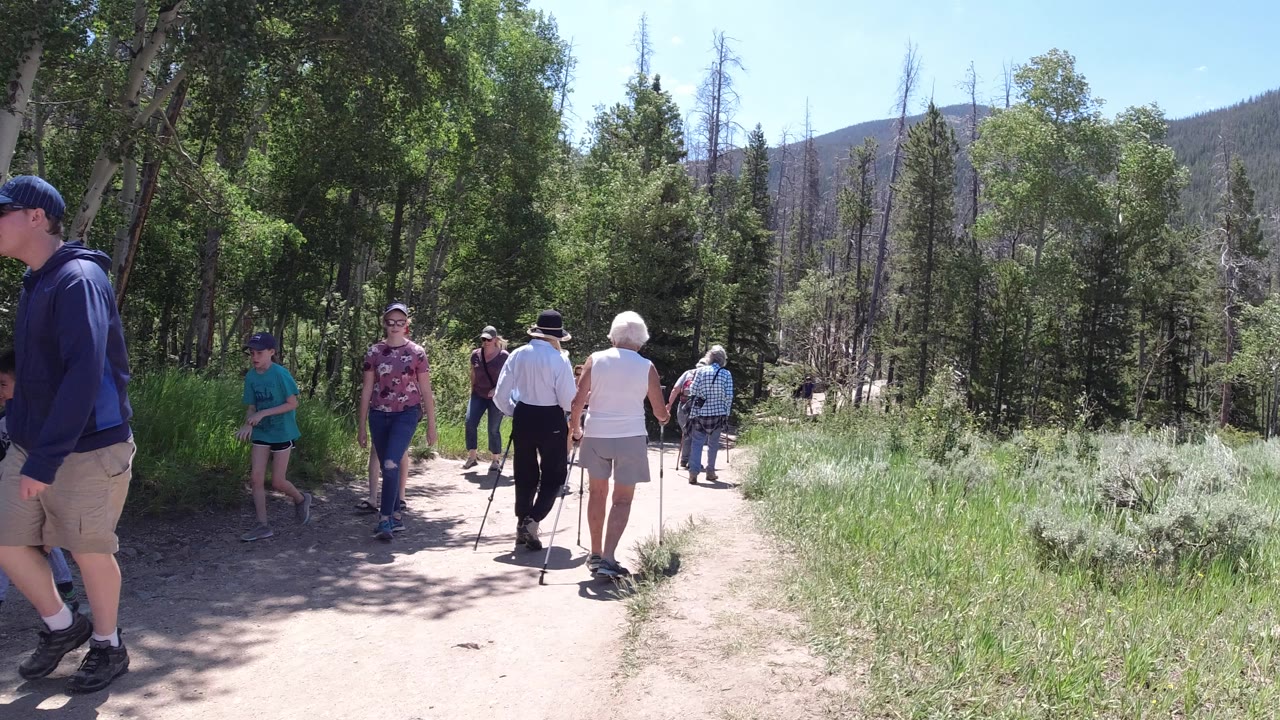 People move in both directions on the Winter Park, Colorado trail, including a child in a teal shirt, adults in sun hats, and older walkers using poles under tall trees.