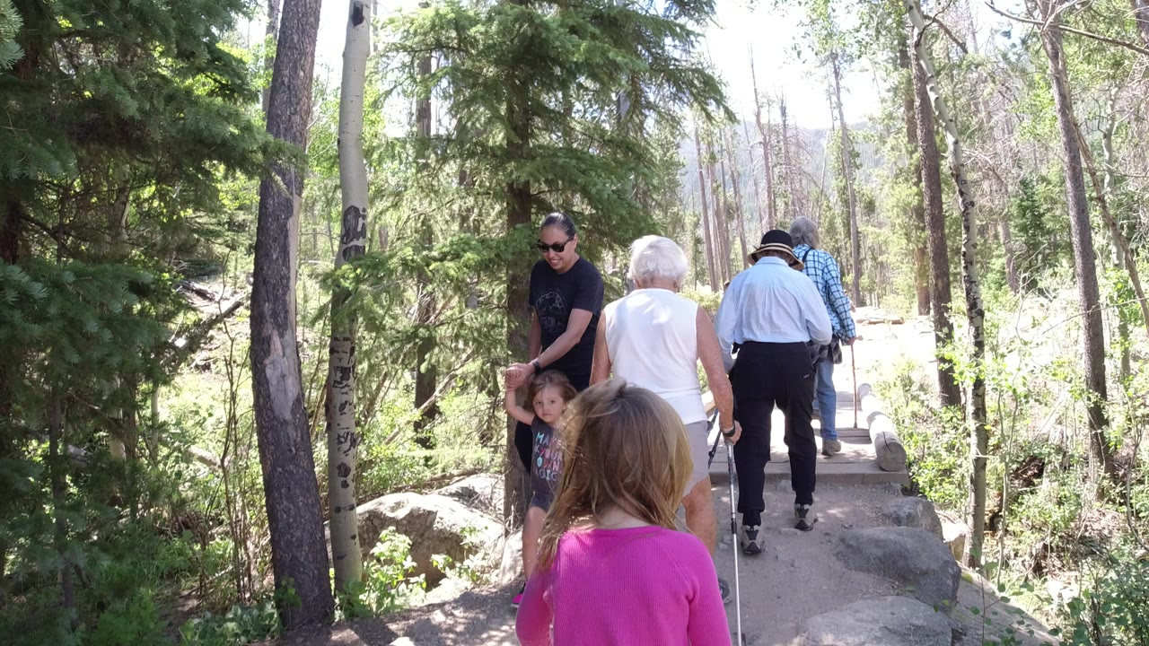 In Winter Park, Colorado, adults and children walk along a narrow wooded boardwalk while an older woman in white moves ahead with walking sticks.