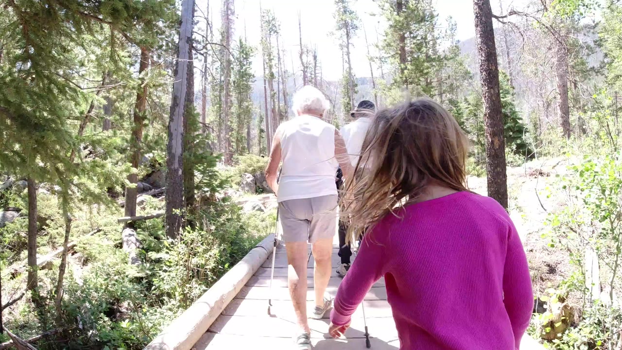 At 0:00 in Winter Park, Colorado, a blonde child in a bright pink top trails two older adults walking away on a narrow wooden path through sunlit trees.