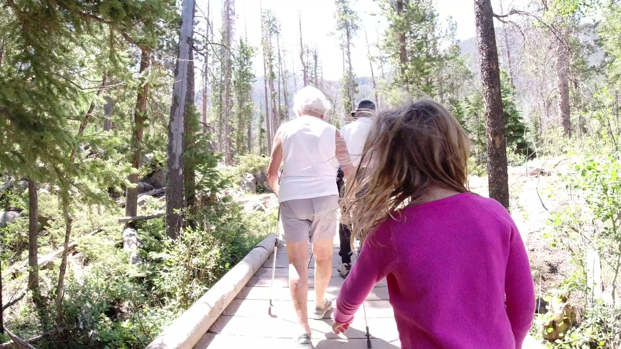 At 0:01 in Winter Park, Colorado, the child remains close to the camera while the older adults continue ahead on the wooden path bordered by shrubs and tree trunks.