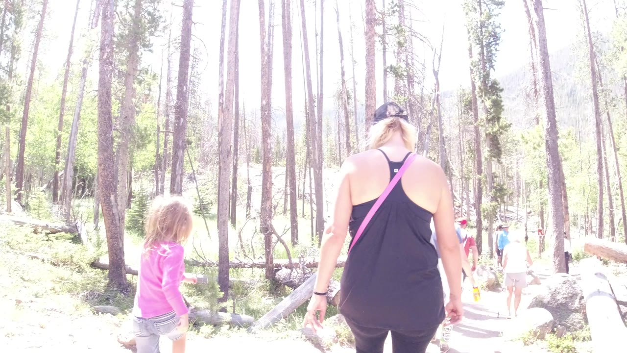At 0:01 in Winter Park, Colorado, the child remains left of frame while the woman walks near center on the sunlit forest steps with hikers farther down the path.