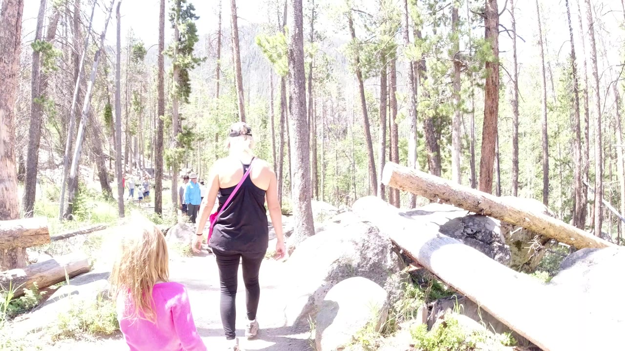 The girl in pink stays near the left foreground while the woman in black walks ahead on the sunlit paved path in Winter Park, Colorado.