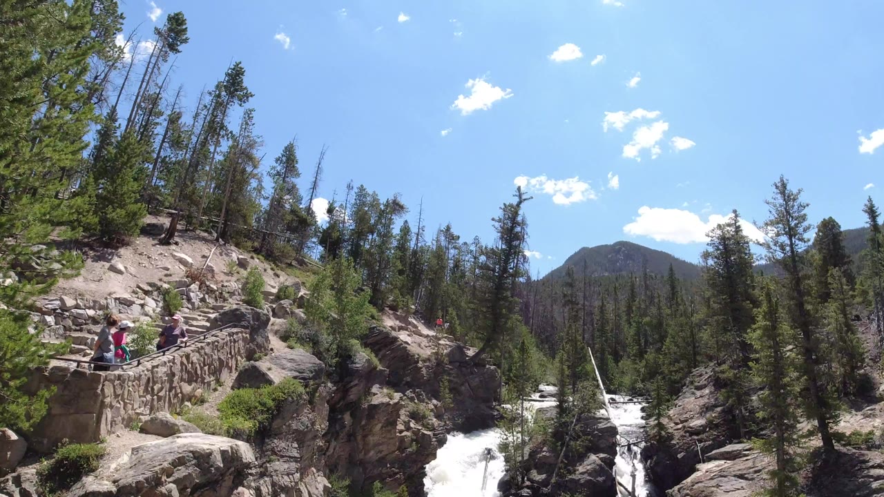 Winter Park, Colorado frame with the camera still wide on the gorge, showing rushing whitewater, the stone overlook at left, conifers, rock ledges, distant mountains, and tiny visitors near the railing.
