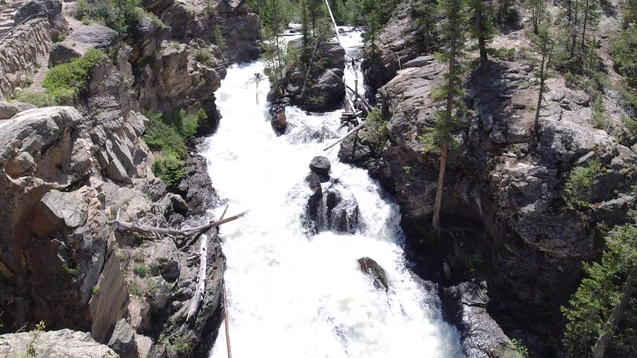 White water surges through a narrow rock channel in Winter Park, Colorado, with pine trees, shrubs, fallen logs, and no visible people.