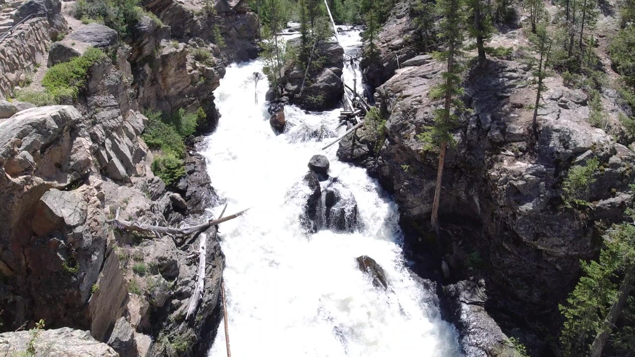 Foaming water drops around dark boulders in a rocky Winter Park channel, with tan cliff faces, scattered trees, and no visible people.