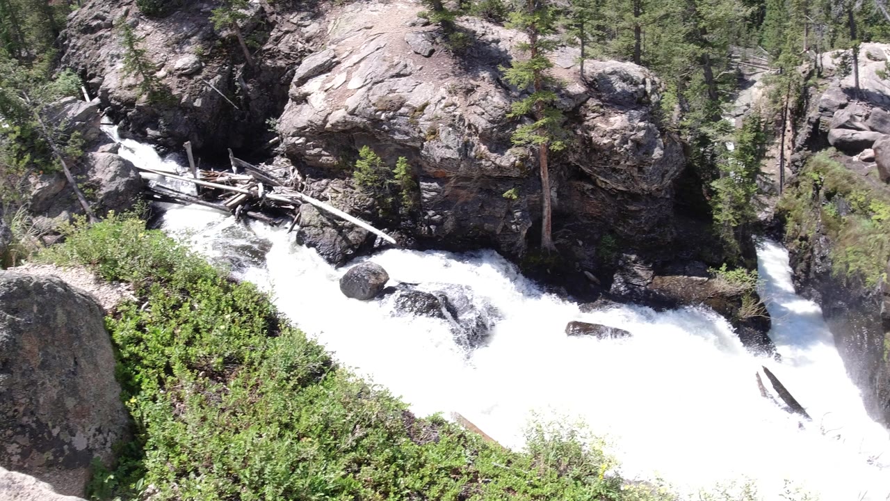 Foaming water runs below a tall rock face in Winter Park, Colorado, with pale fallen logs across the channel, dense greenery, and no visible people.