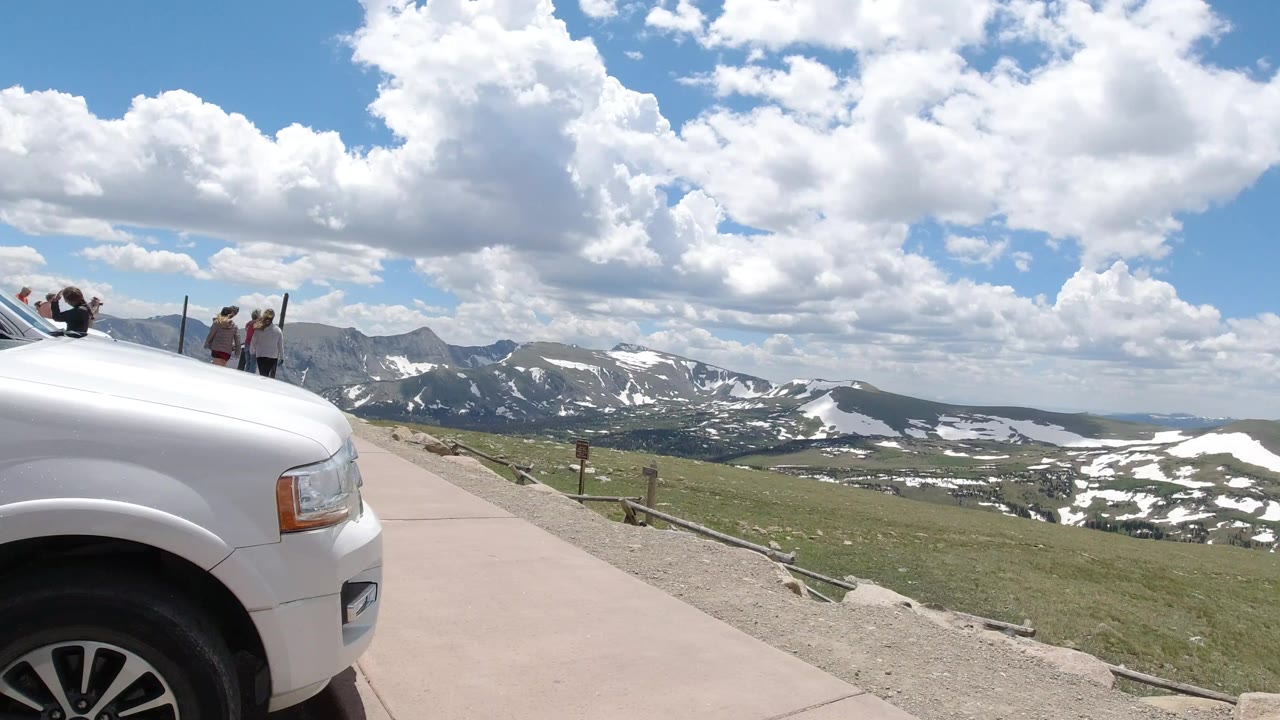 White vehicle at a mountain overlook with distant snow patches and clouds.