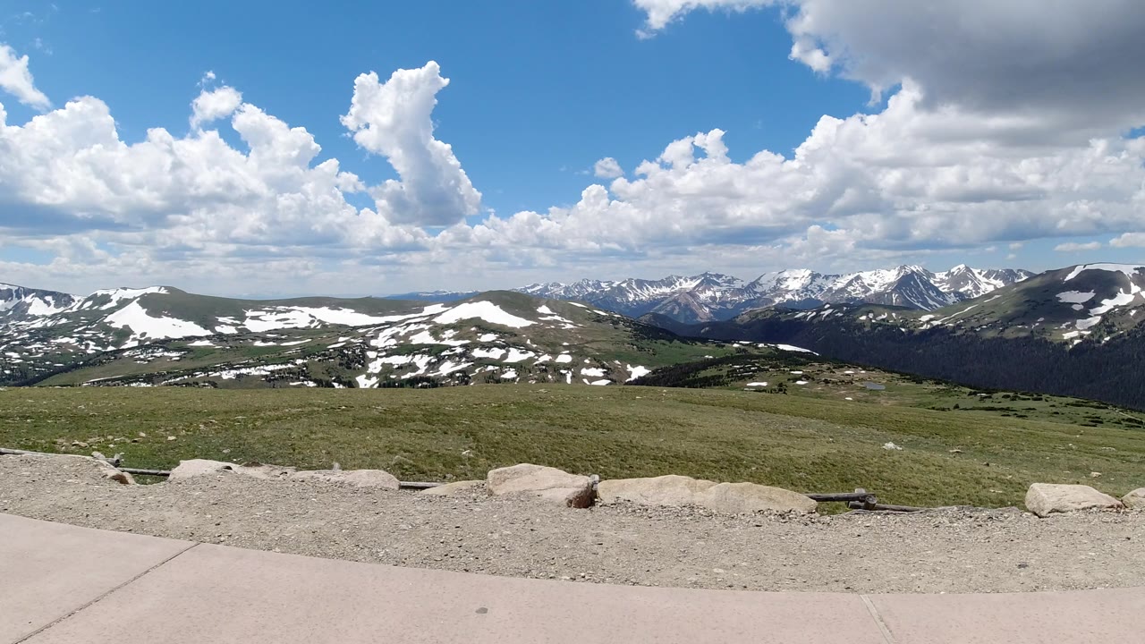 Wide mountain overlook with sidewalk, grass, snow patches, and clouds.