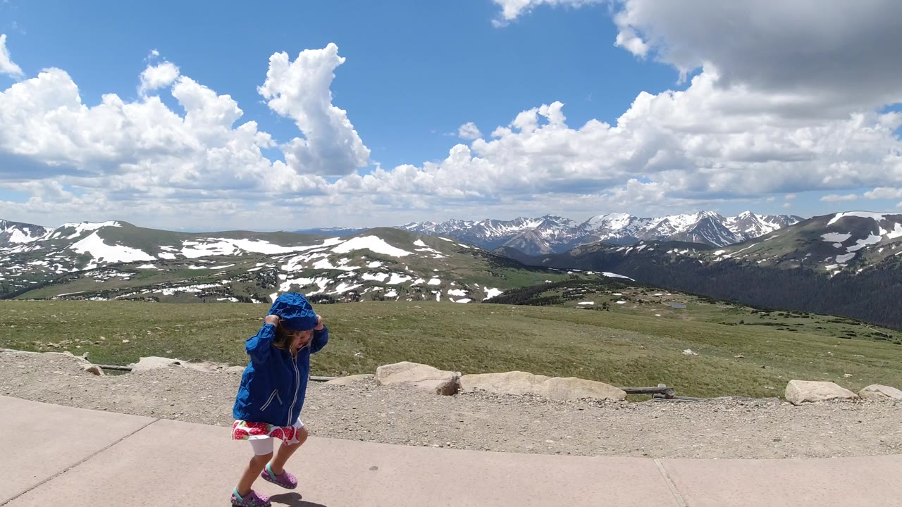 A child in a blue hooded jacket walks along a paved sidewalk at a Winter Park, Colorado mountain overlook, with grassy slopes, snow patches, and distant peaks under bright clouds.