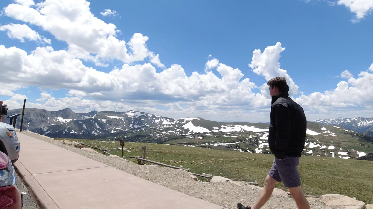 Man walking beside a mountain overlook path under bright clouds