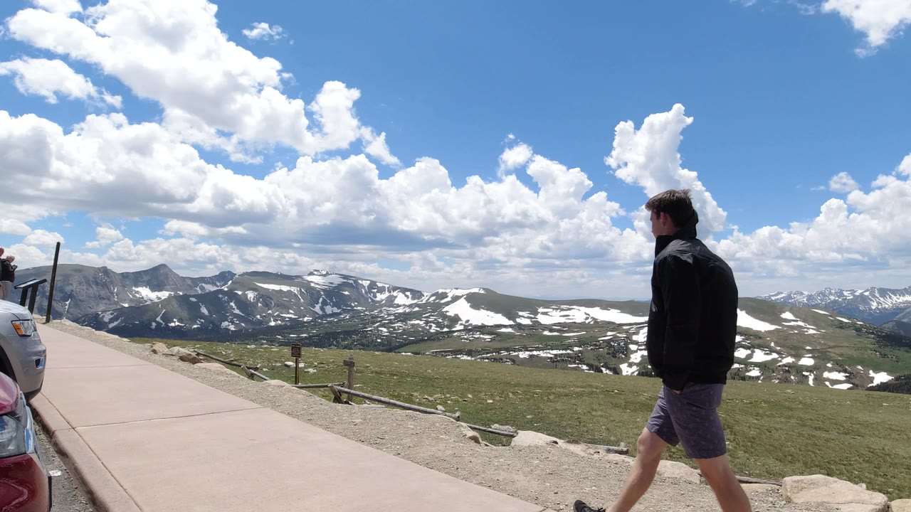 The man continues along the paved path near the right side of the Winter Park, Colorado frame, while snow-streaked ridges and a cloud-filled blue sky fill the distance.