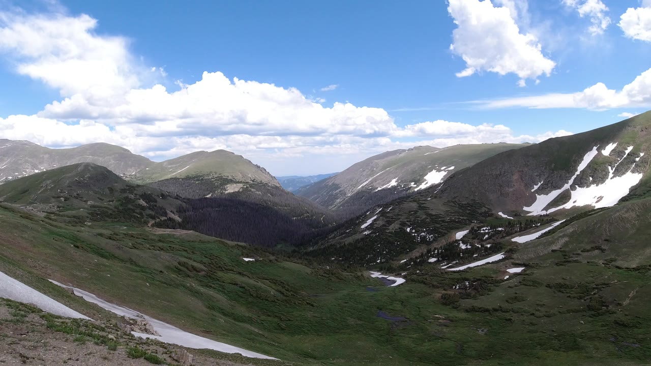Wide Winter Park, Colorado mountain valley with green slopes, snow patches on the right ridge, bright clouds, blue sky, and no visible people.