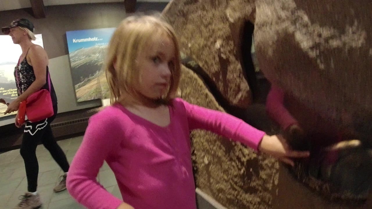 Young girl in a pink top stands close to a large reptile enclosure in Winter Park, Colorado, with an adult visitor and wall sign behind her.