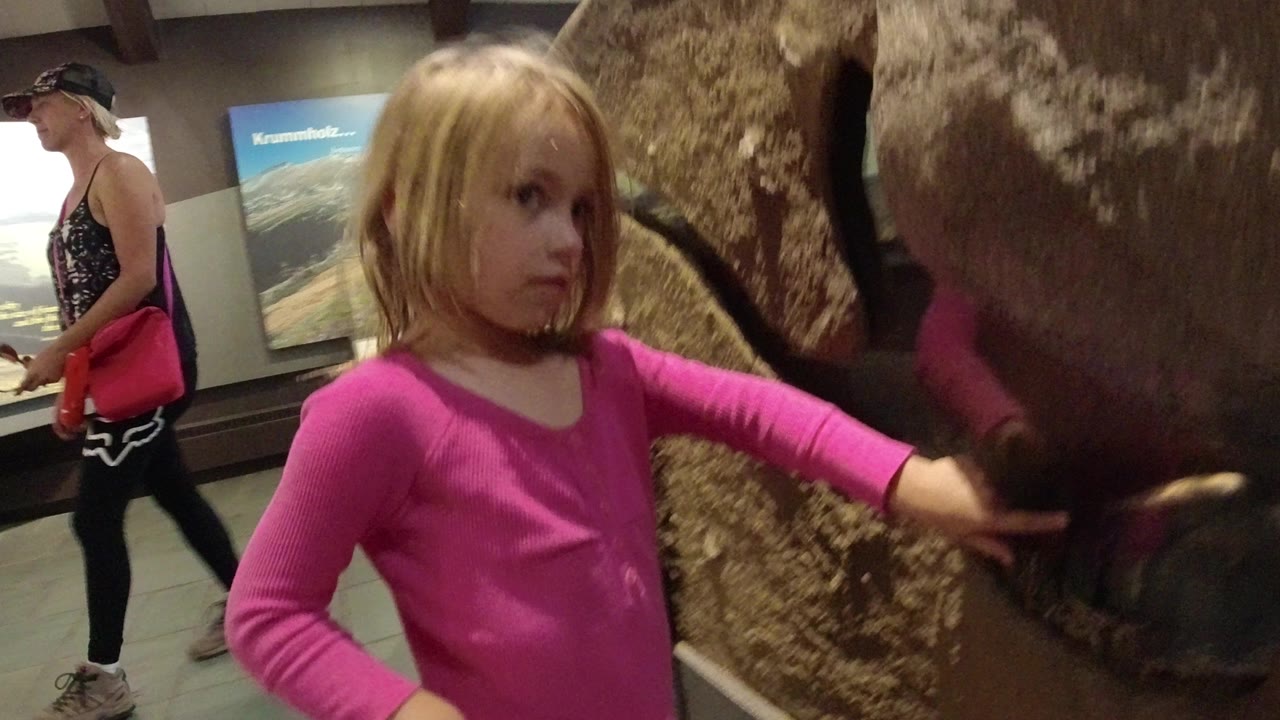 The girl faces the enclosure glass in Winter Park, Colorado, while the large brown reptile form fills the right side of the frame and a visitor remains in the background.
