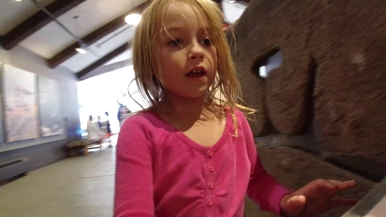 Blonde girl in pink shirt near a rough rock exhibit inside a museum.