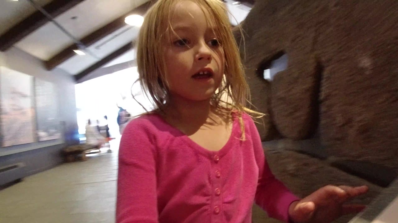 The girl remains close to the lens in the museum, with her pink shirt prominent and a dark textured rock wall or large sample filling the right side of the frame.