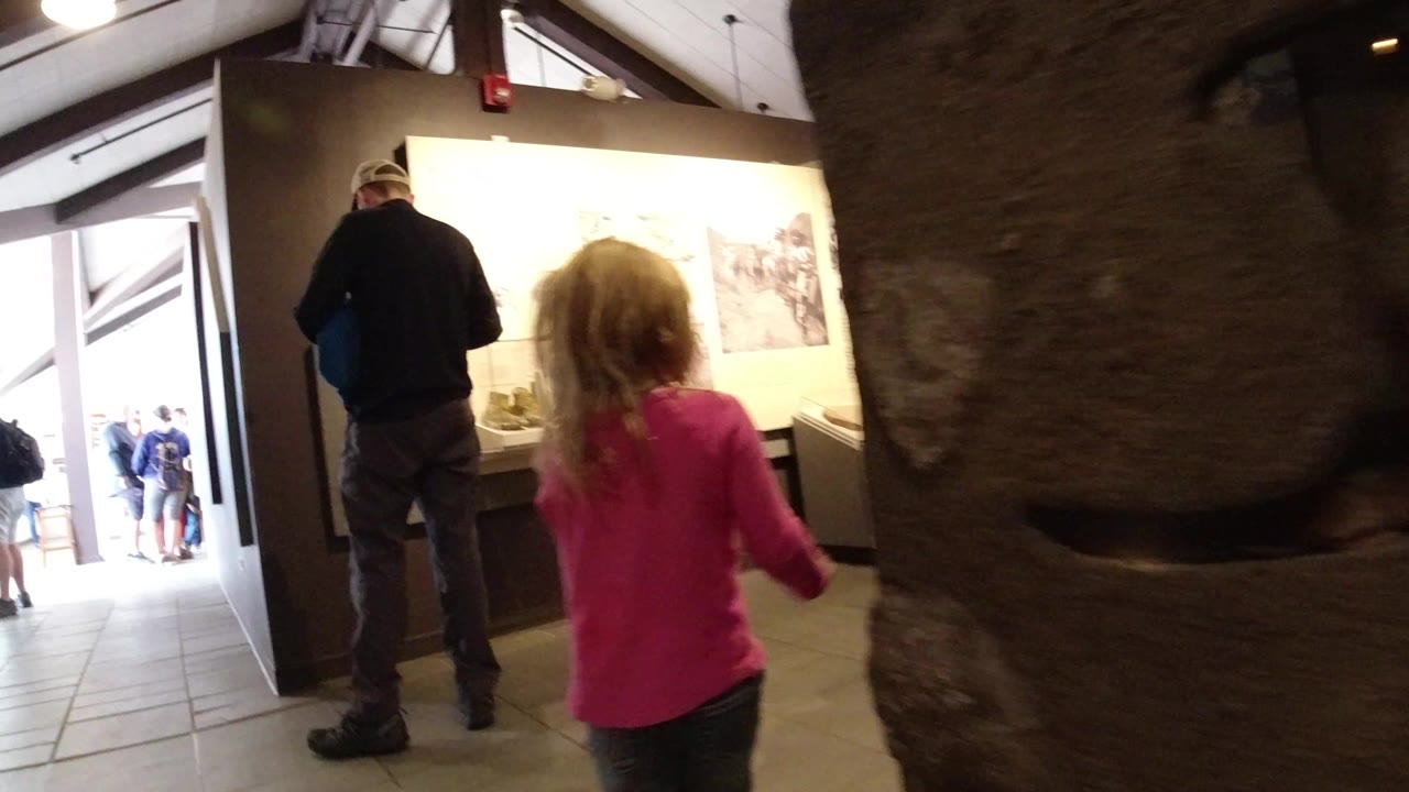 In Winter Park, Colorado, a child in a bright pink shirt walks into a warm-lit museum gallery with adults ahead, wall displays on the right, and a large rock-textured structure near the center.