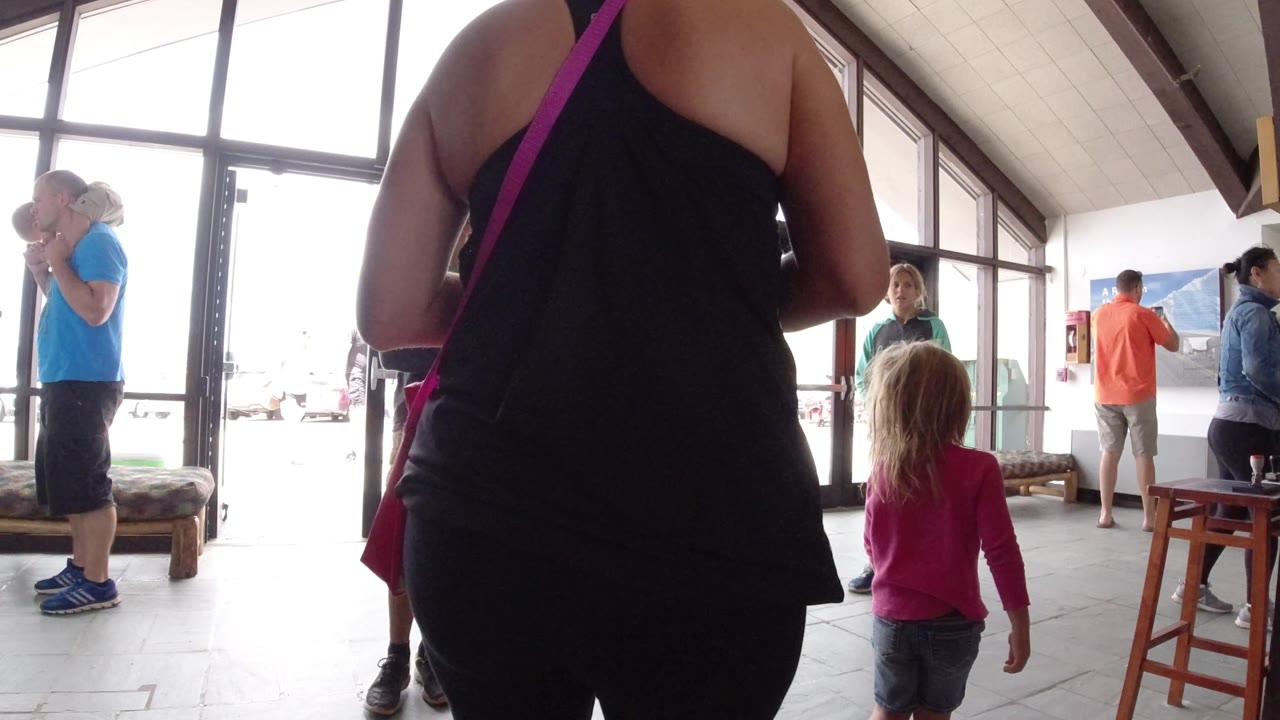 A young girl in a pink shirt faces a wooden table in a Winter Park, Colorado interior while an older woman stands close in the foreground and visitors remain near the glass entrance.