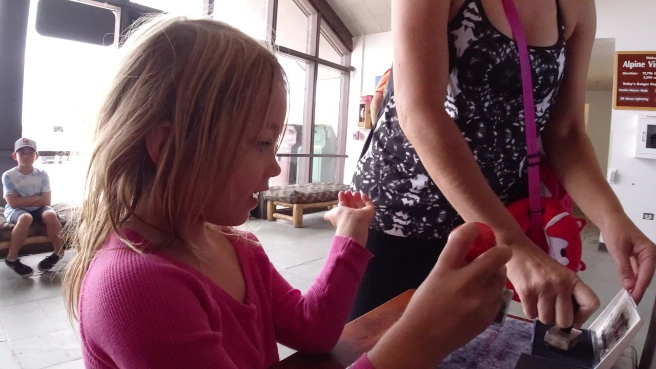 Young girl in a pink sweater holding a stamp beside an adult at a table.