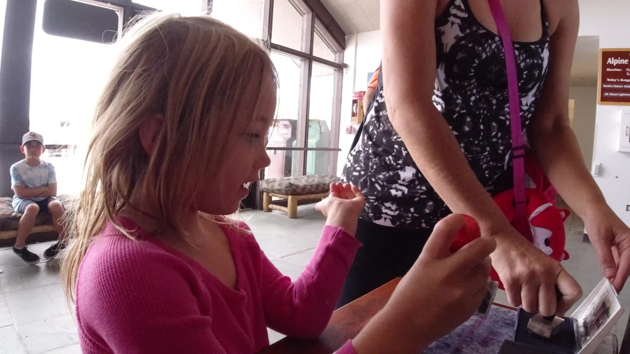 In Winter Park, Colorado, the girl looks toward the tabletop as she positions the red-handled stamp near an adult's hand, with a seated person and bright doors behind her.